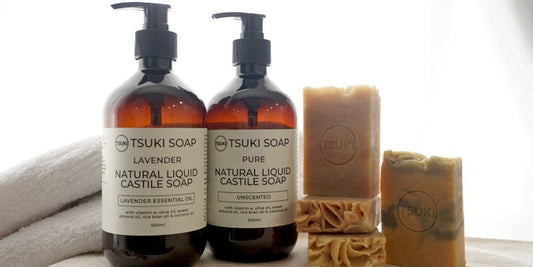 2 bottles of Tsuki natural handmade liquid castile soap, lavender and pure unscented, bars of Tsuki Soap's natural handmade soap stacked on the right side.